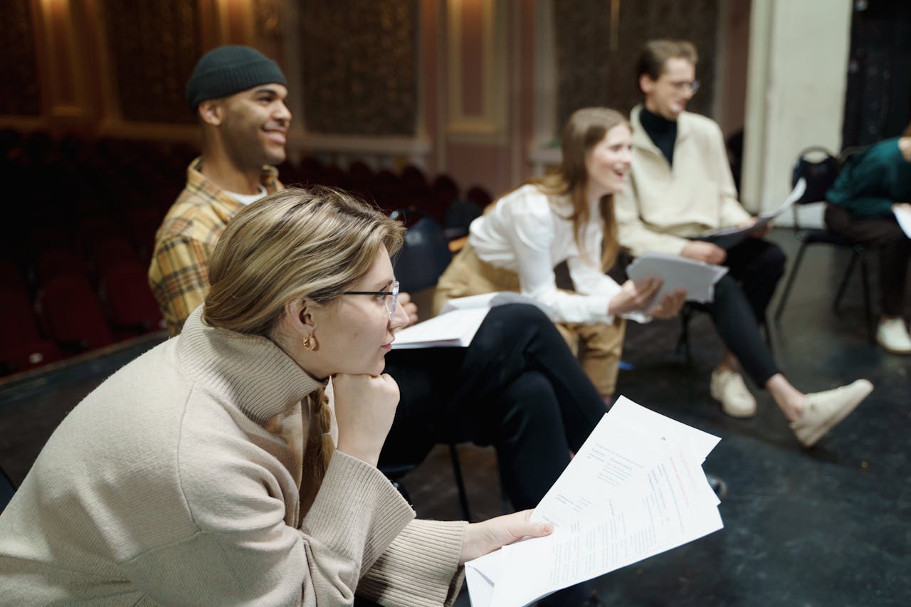 Actors rehearsing a play on stage, holding scripts and discussing roles in a theater setting.